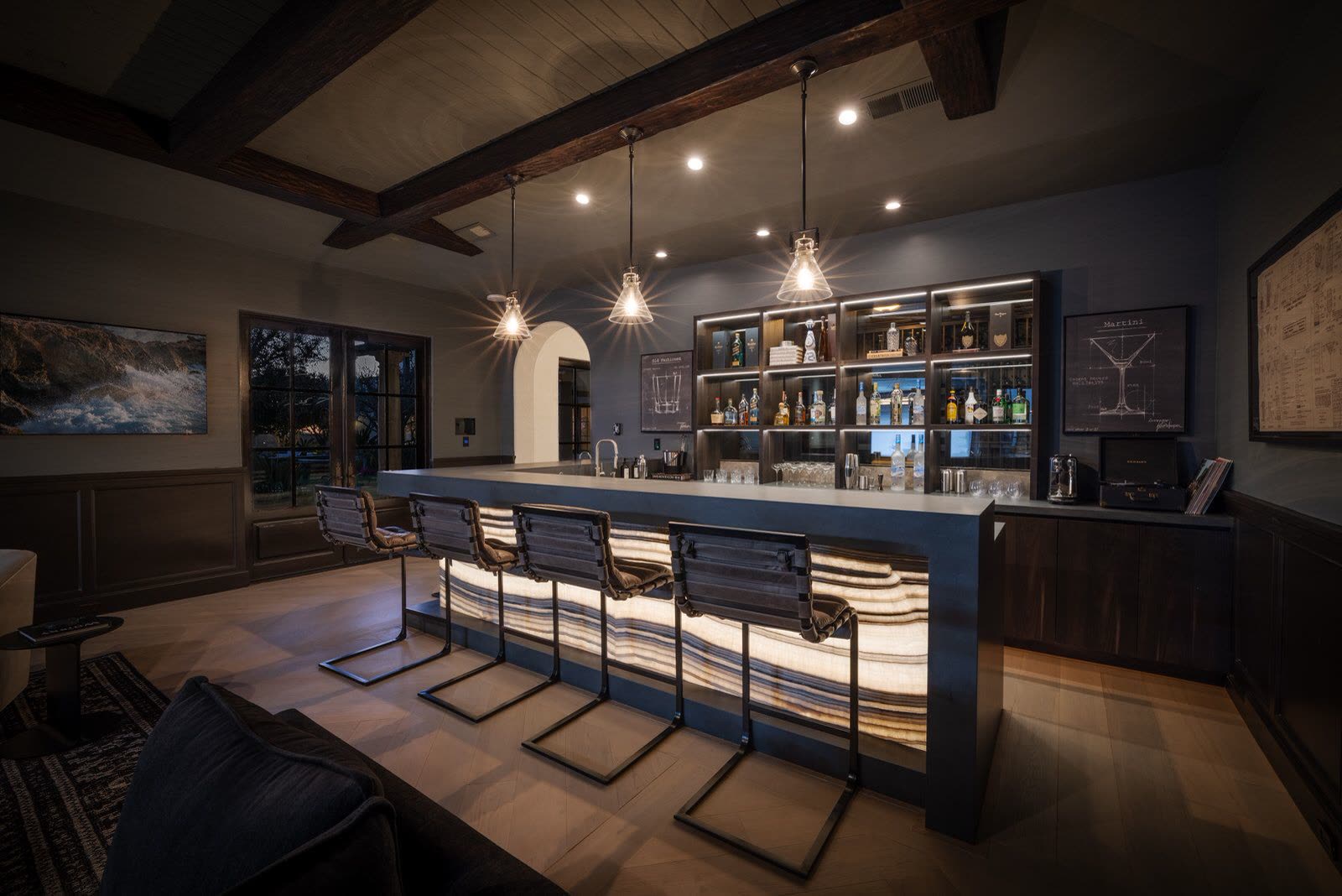 Home bar with a backlit onyx counter front, dark timber beams and glass shelving