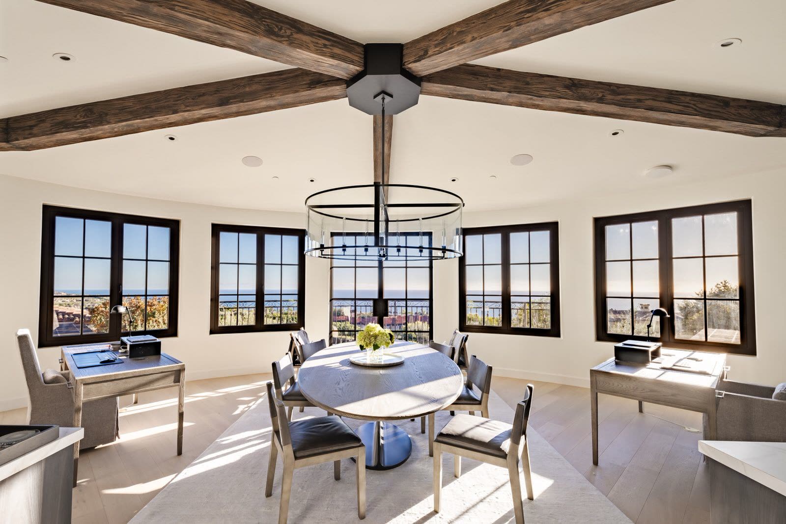 Round dining table beneath a ring chandelier, framed by a wall of black-framed windows